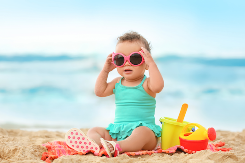 A baby sitting on the beach wearing sunglasses.