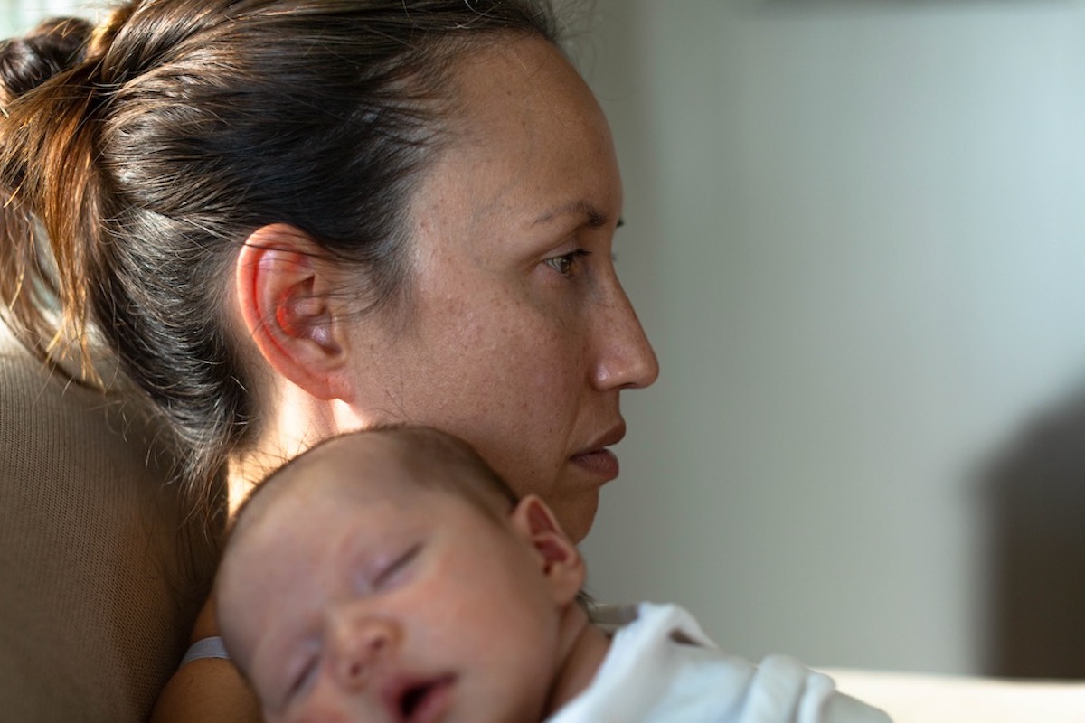 An unhappy-looking mom holding her baby at home sitting on the couch thinking and looking off into space.