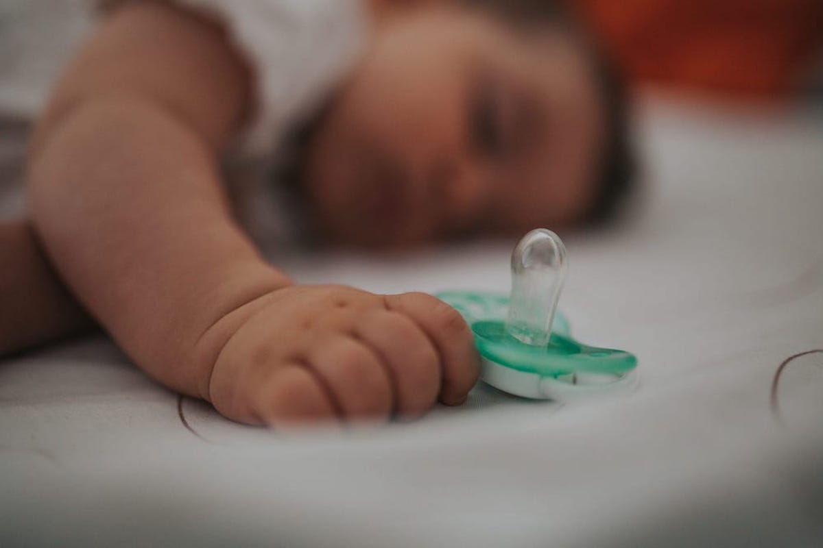 An infant sleeps on his stomach in a crib, gripping a green pacifier in his hand.