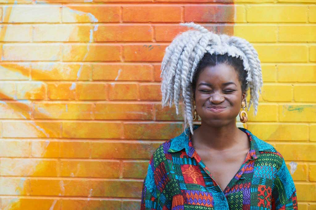 A person stands against a yellow wall and holds their breath while smiling.