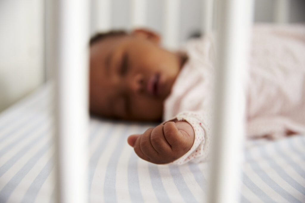 An infant wearing pink sleeps on her back in a crib with arms outstretched.