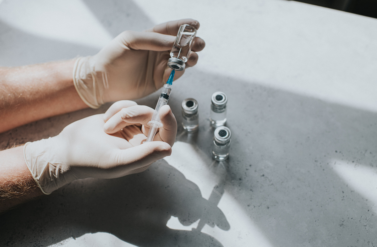 A hand wearing latex gloves holds a vial of clear liquid and a syringe.