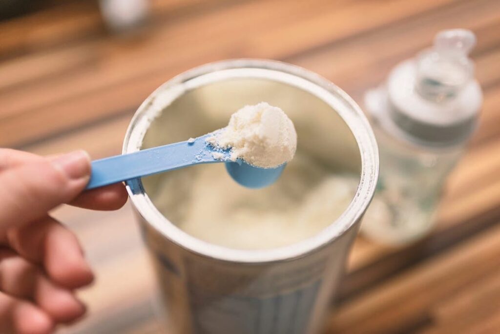 A canister of baby formula is pictured with a hand holding a full scoop.