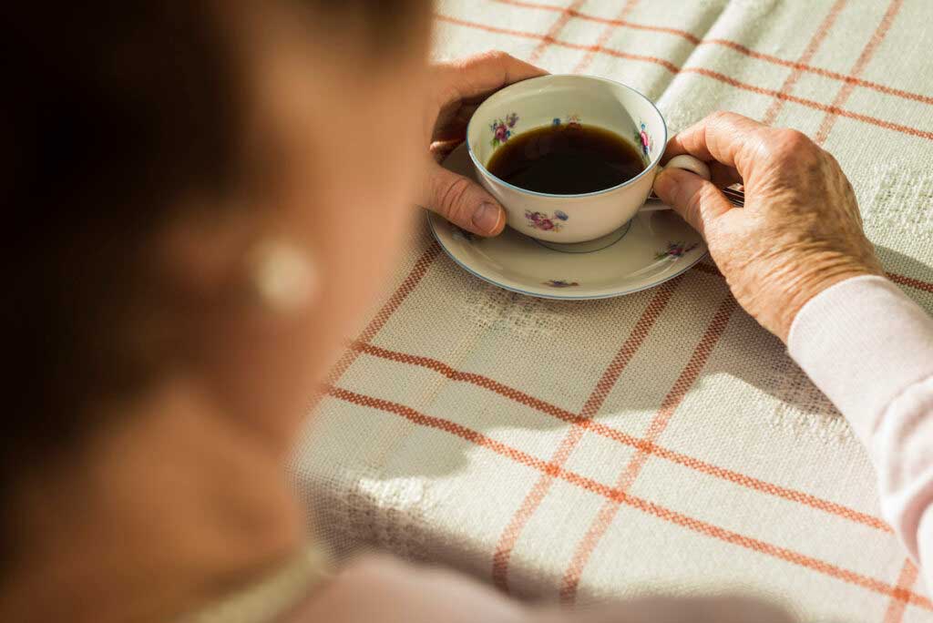 A senior woman is seen from behind, sitting at a table and drinking a cup of coffee.