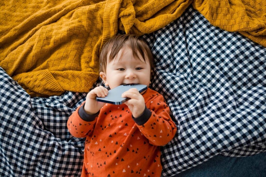 A small child reclines on picnic blankets and chews on a cellphone while smiling.