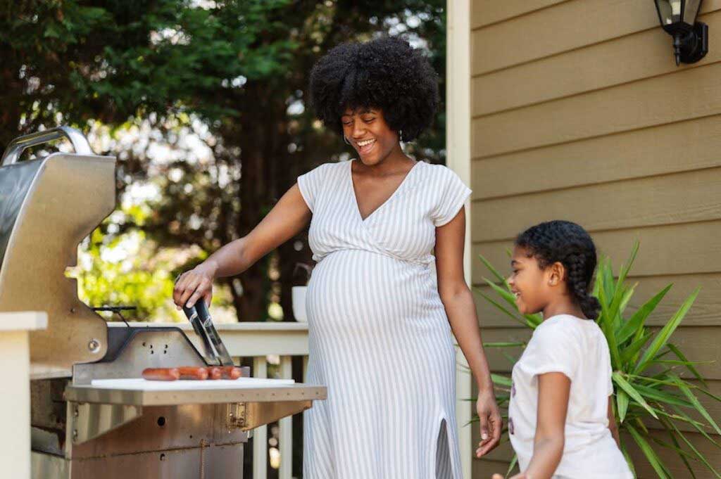 A pregnant woman wearing a white dress stands over a grill preparing hotdogs while a child looks on.