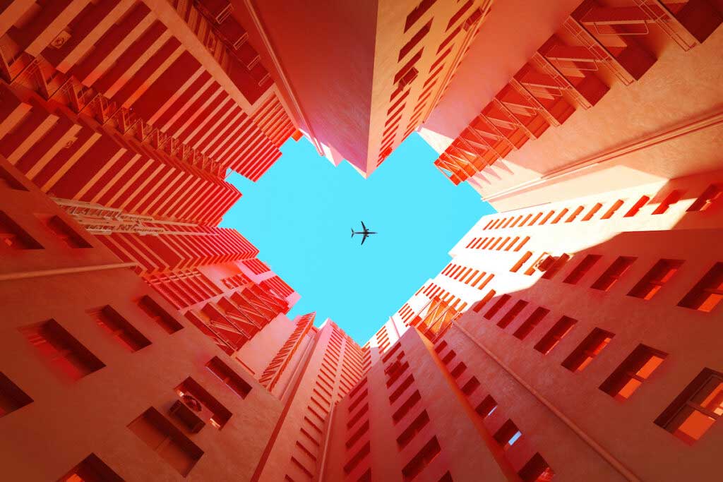 A plane flies through a patch of blue sky in between orange buildings.