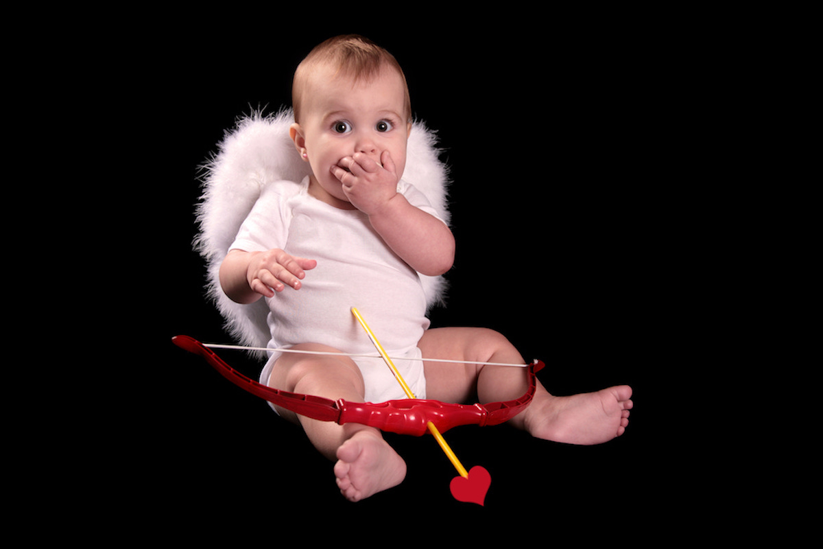 A baby is seated against a black background wearing feather wings and holding a red cupid's bow.