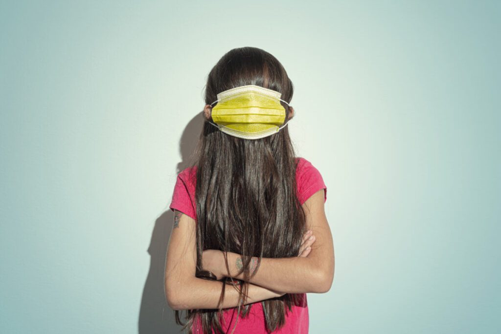 A girl stands with her hair over her face and a mask over her hair, with arms crossed, in front of a blue background.