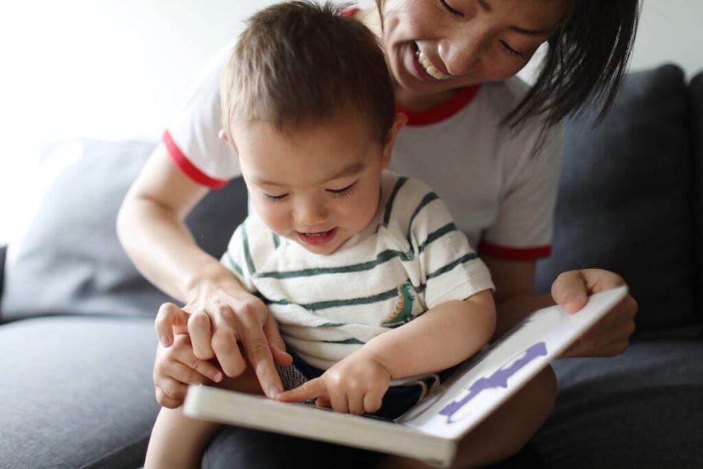 A mother holds a toddler on her lap and, together, they point at images in a book.