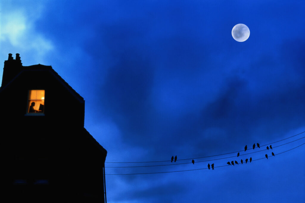 A window in a house is lit up in the night with a full moon.