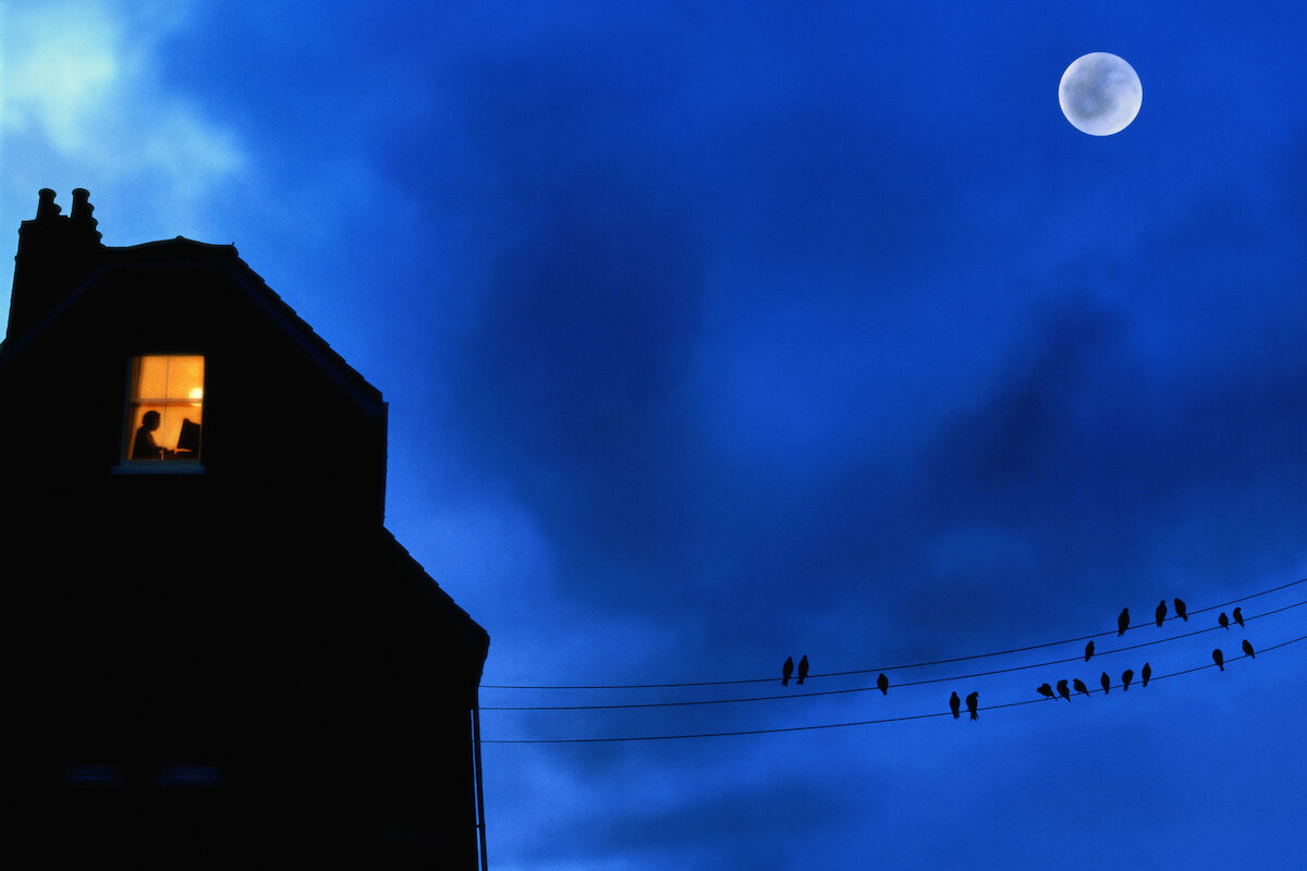 A window in a house is lit up in the night with a full moon.