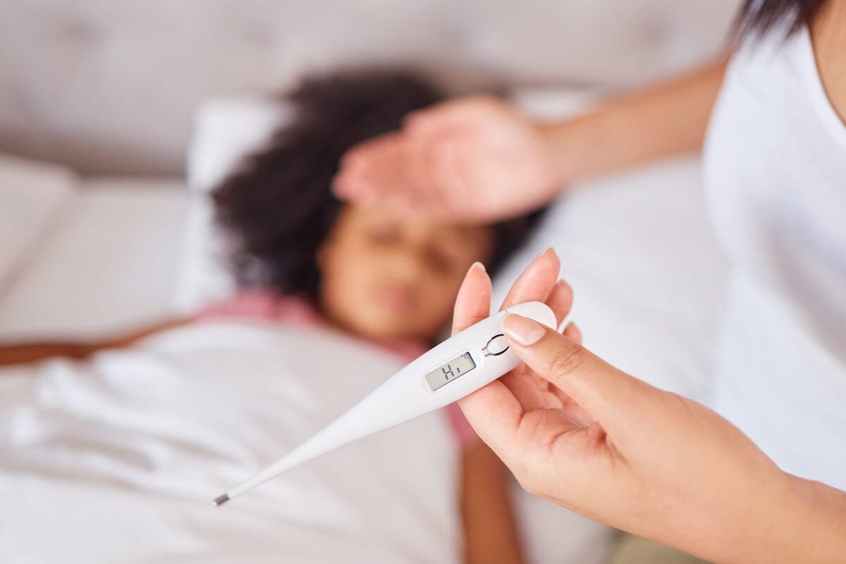 A parent holds the back of her palm to her child's forehead while the child sleeps in bed. In the other hand, the parent holds a thermometer that reads "hi"