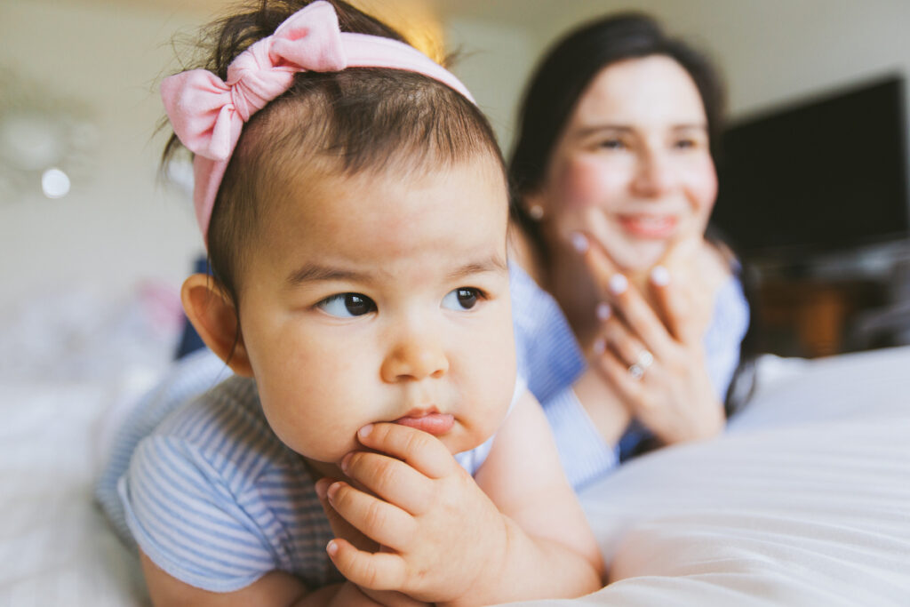 A skeptical-looking infant lies on a bed with a parent.