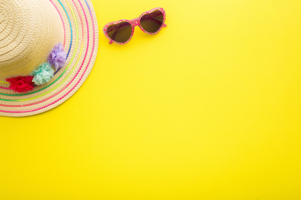 A straw infant hat and pink heart-shaped sunglasses are seen on a yellow background.