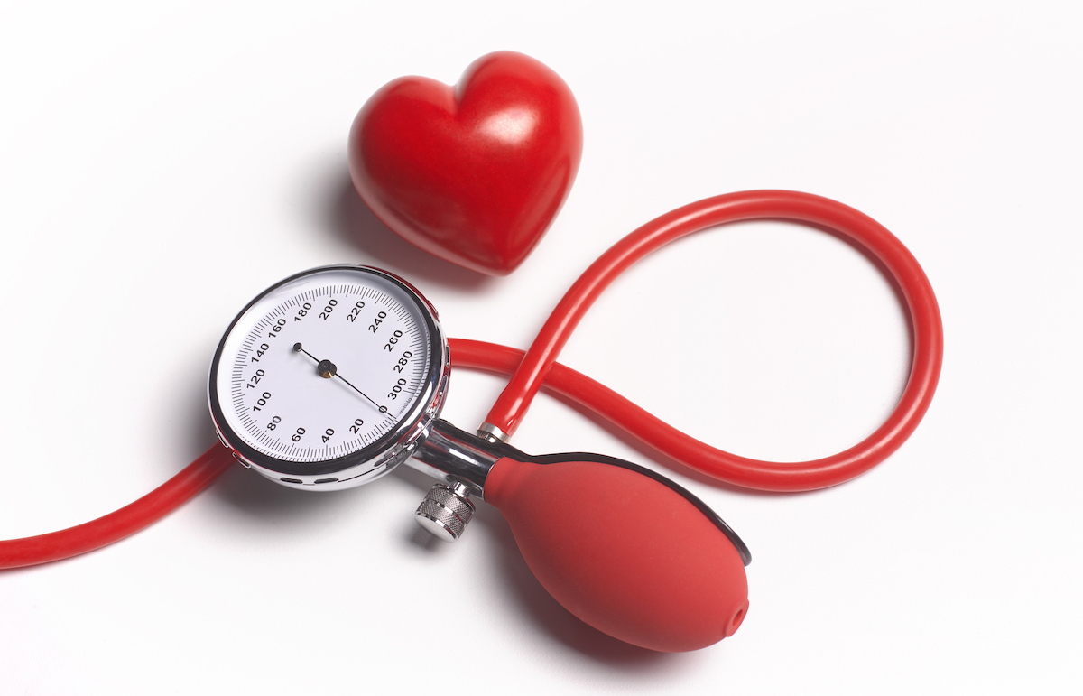 A red manual blood pressure gauge and a plastic red heart are seen on a red background.