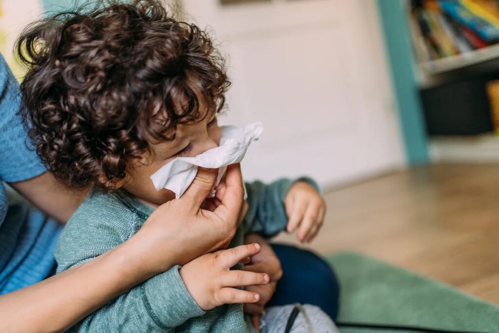 A toddler is seen sitting on the lap of a parent. The child is blowing their nose into a tissue held by the parent.