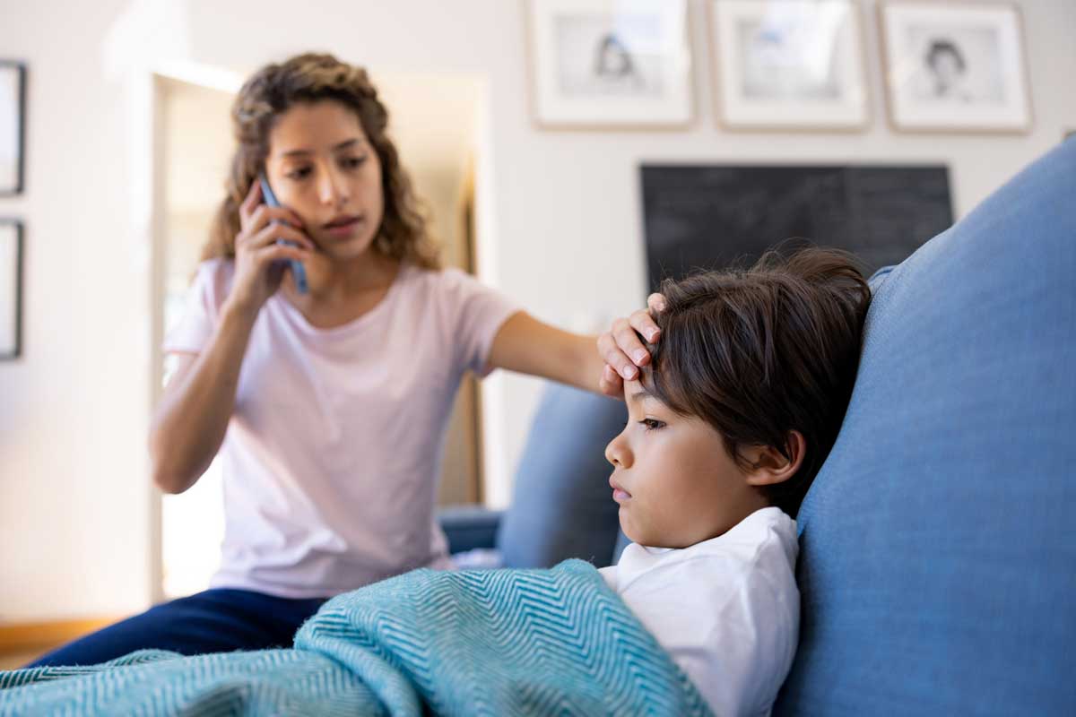 A parent looks worried as she talks to a doctor on the phone while placing her palm on the forehead of a sick child.