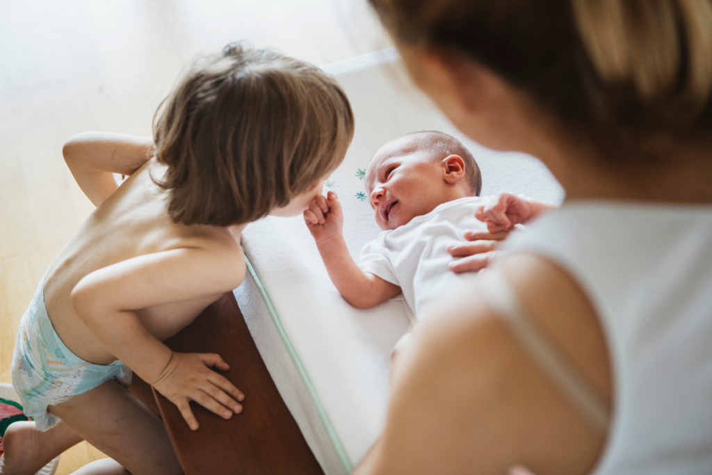 A toddler looks on as their parent changes a new sibling's diapers.