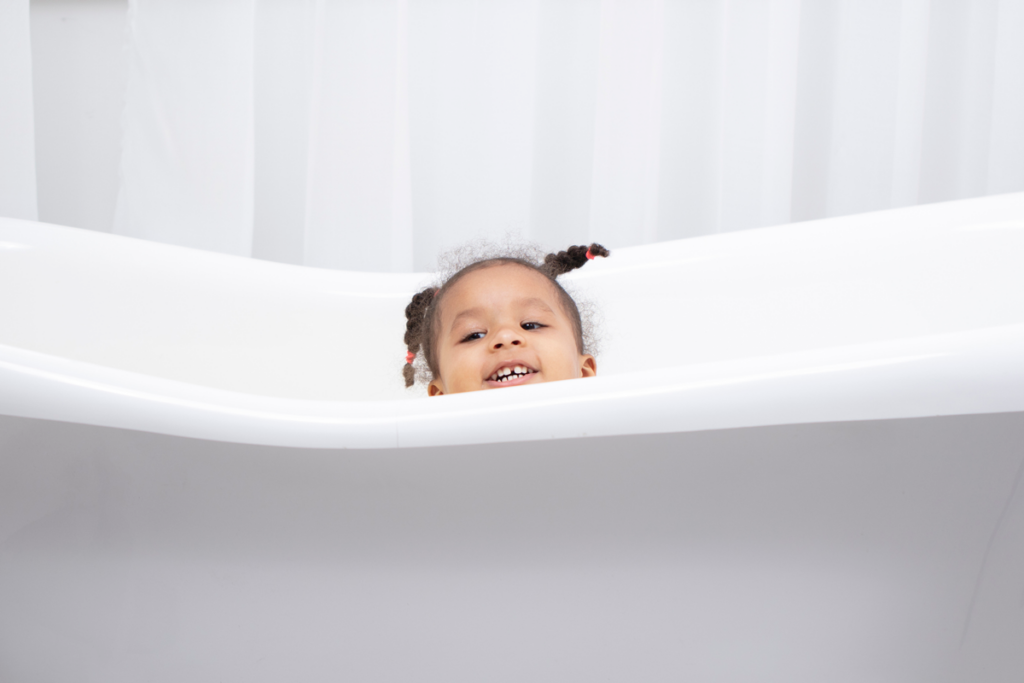 A small child smiles as they peep over the side of a white bathtub.