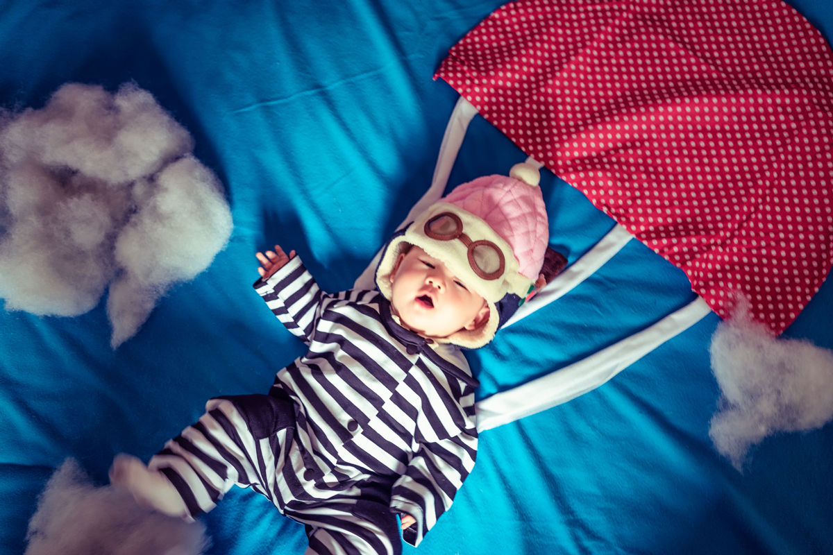 A baby on a blue sheet wears a pretend parachute and skydiving goggles.