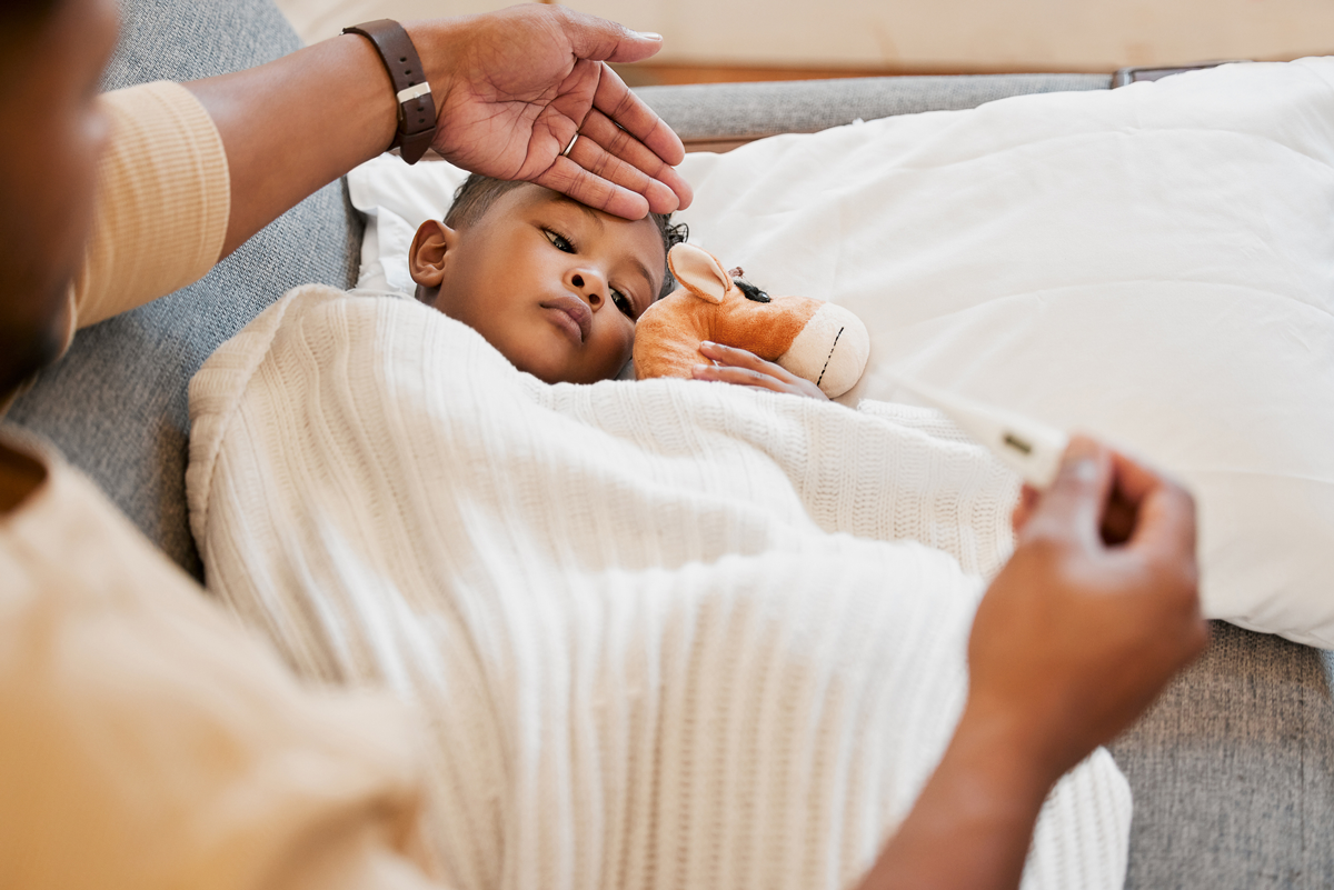 A parent places their hand on their baby's forehead to test for a fever. The baby is tucked under a blanket and looks sick.