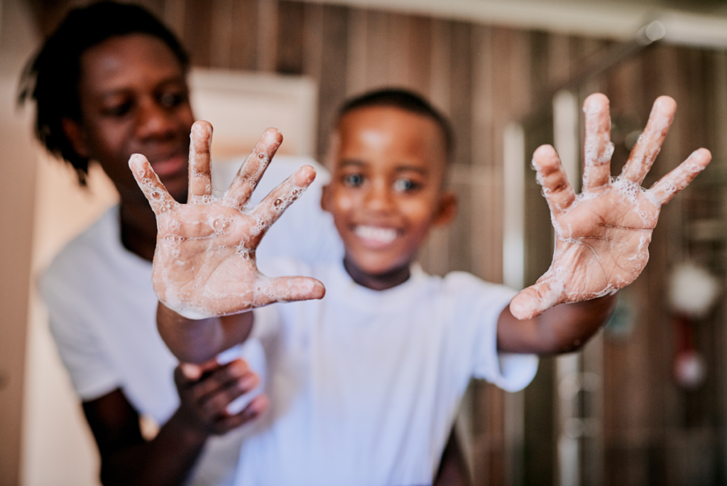 A small child smiles and holds up bubble-covered hands while washing their hands.