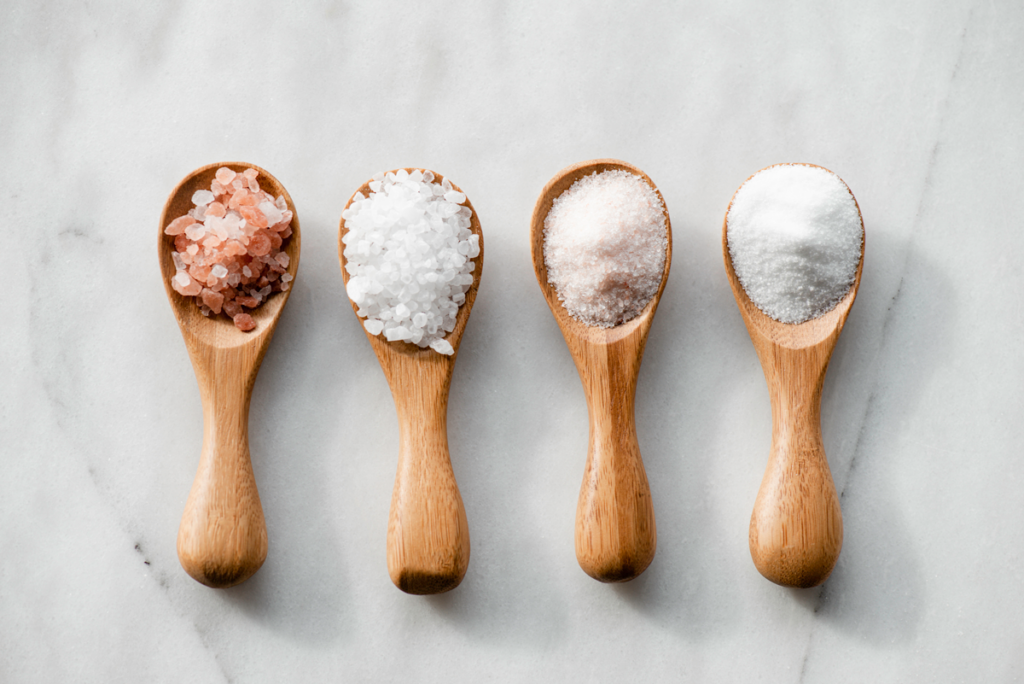 Four tablespoons of various kinds of salt are pictured on a marble background.