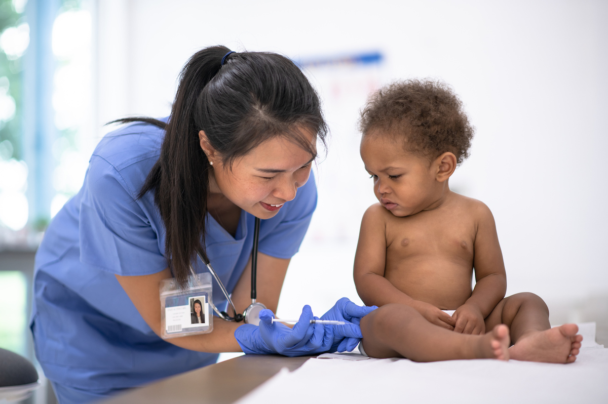 A baby sits on a doctors table and looks concerned while receiving a vaccination from a doctor.