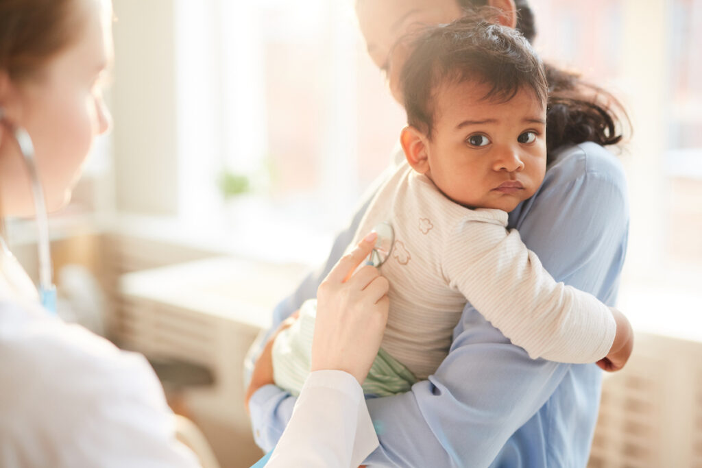 A parent holds and infant while a doctor listens to their lungs.