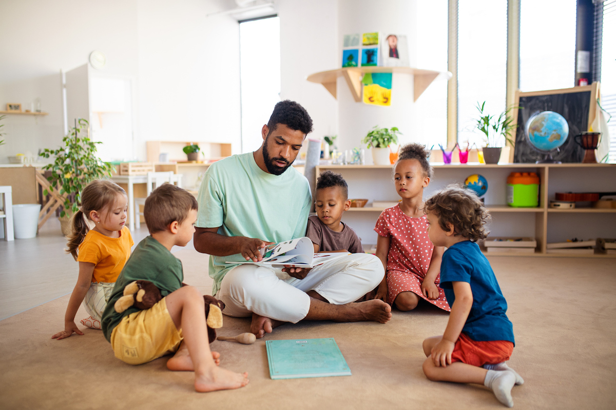 A small group of children sits attentively around their teacher, who is reading them a book.