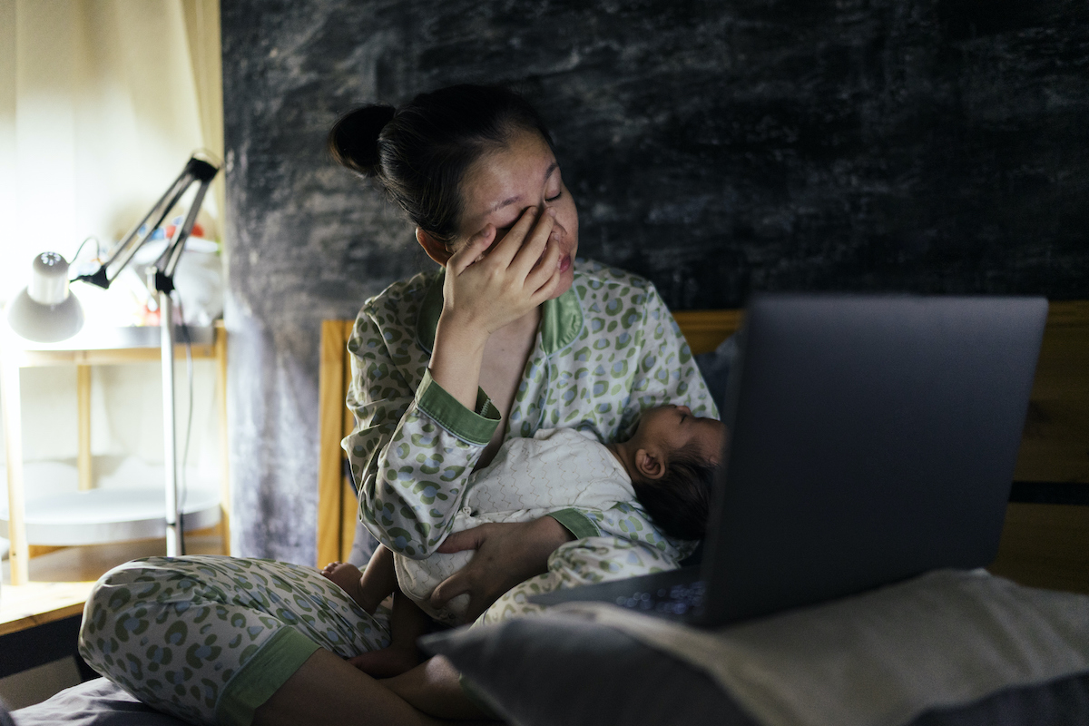 A parent holding a baby on a bed wipes their eyes from exhaustion.