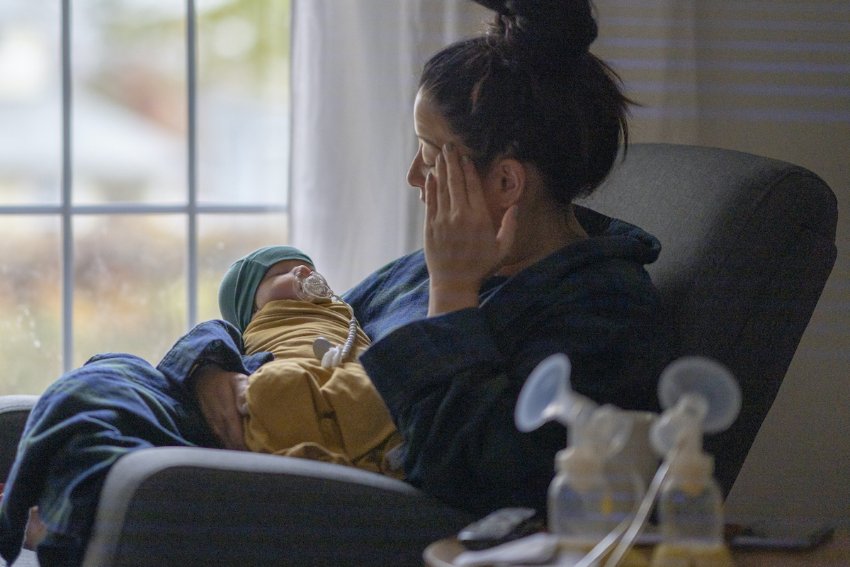 A parent holds their head while holding a baby with a breast pump nearby.