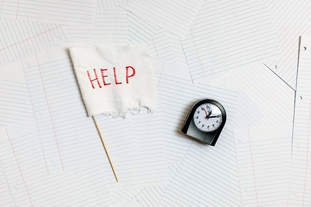 Black alarm clock laying on a pile of lined sheet paper next to a white surrender sign that says HELP in red ink.