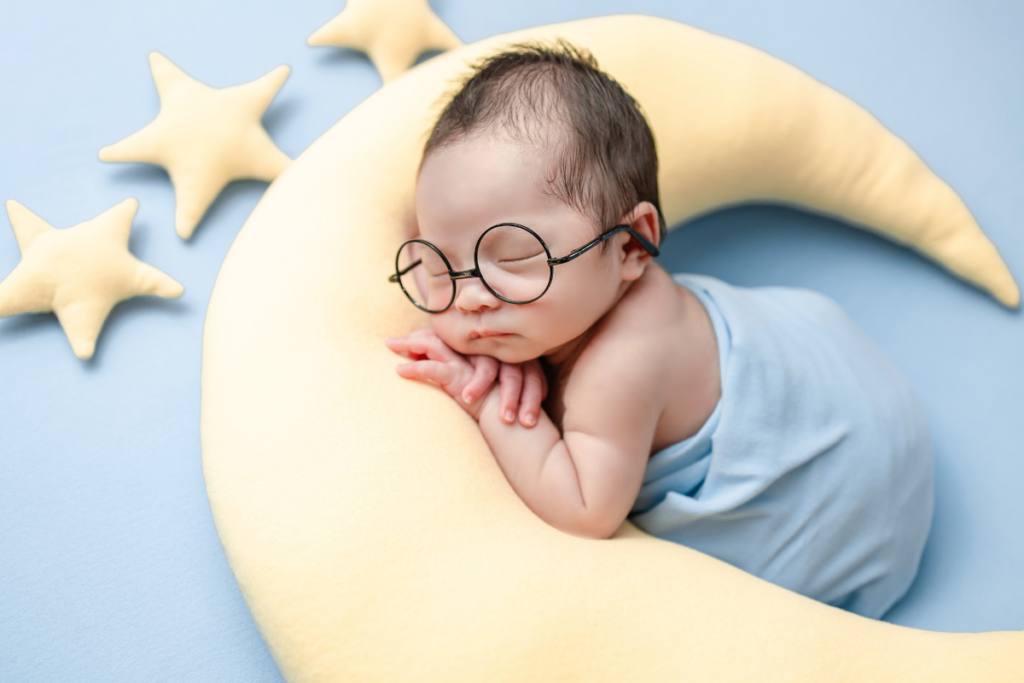 A baby wearing glasses and sleeping on top of a moon shaped pillow.