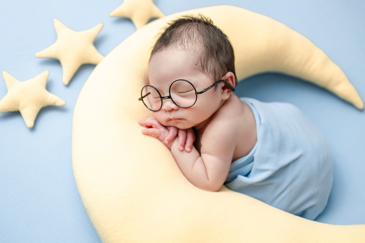 A baby wearing glasses and sleeping on top of a moon shaped pillow.