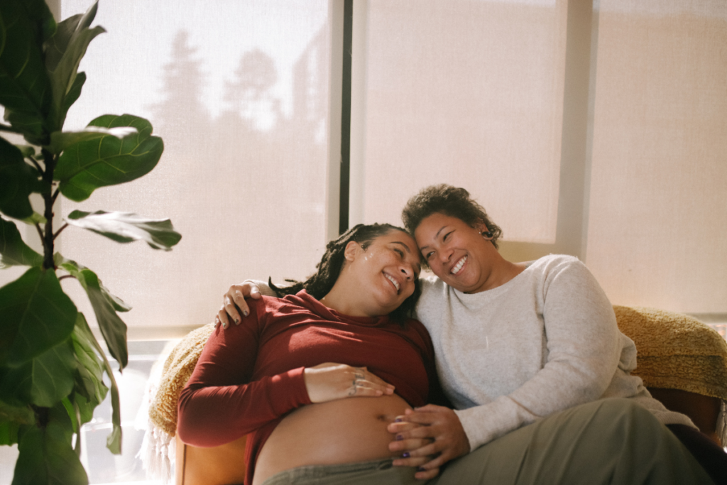 Two partners, where one of them is pregnant sitting on the couch embracing each other