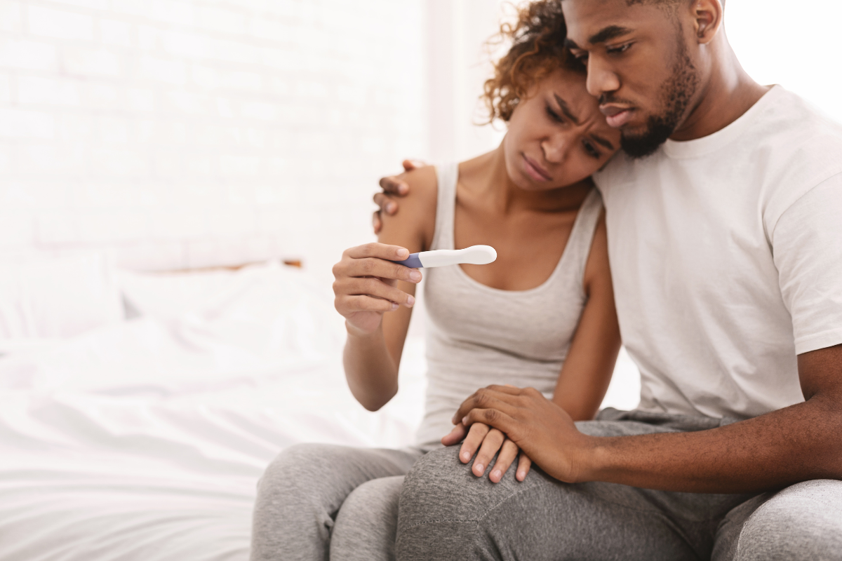 A couple sitting on a bed and looking at a pregnancy test disappointed.
