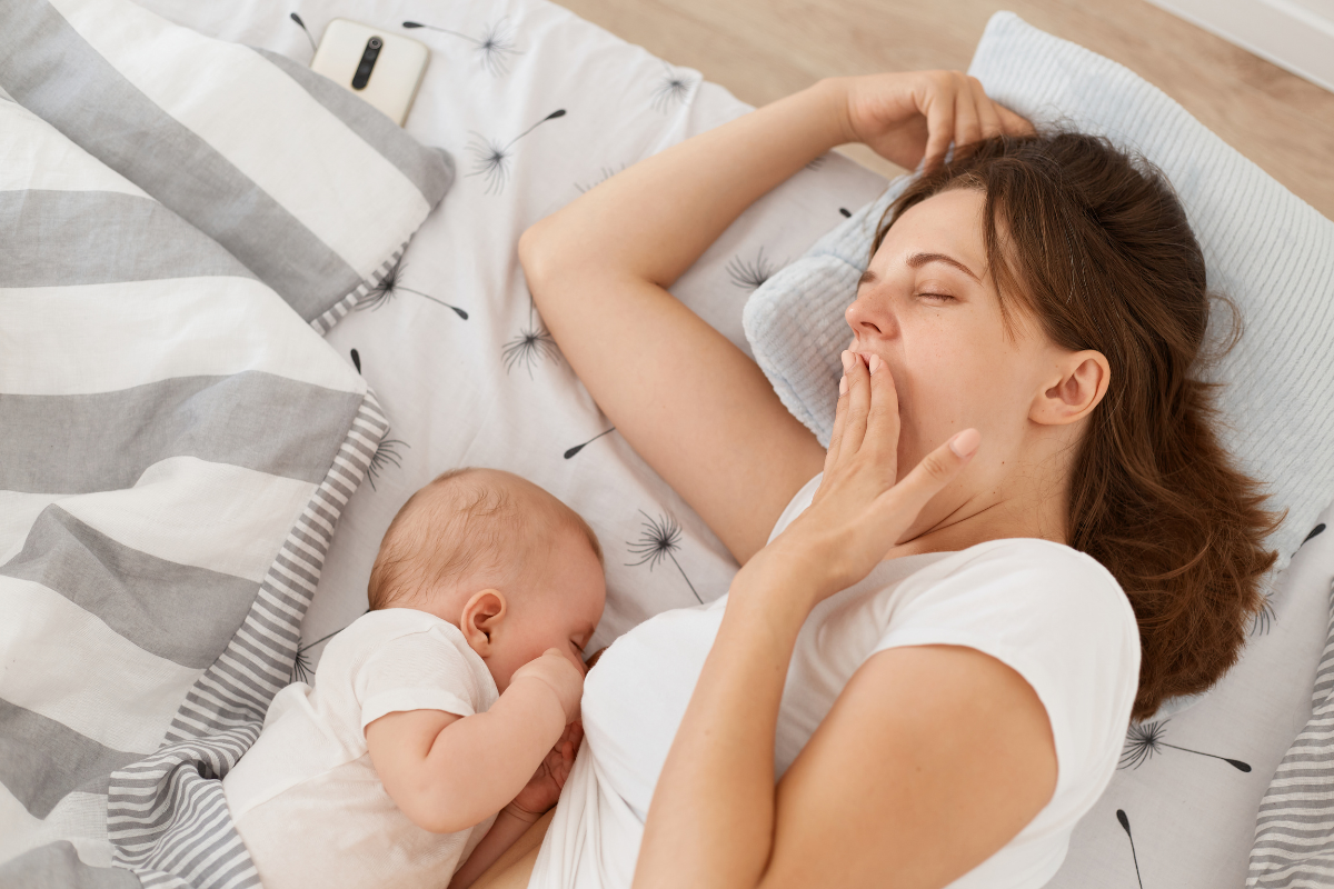 A parent yawning while breastfeeding her baby in bed.