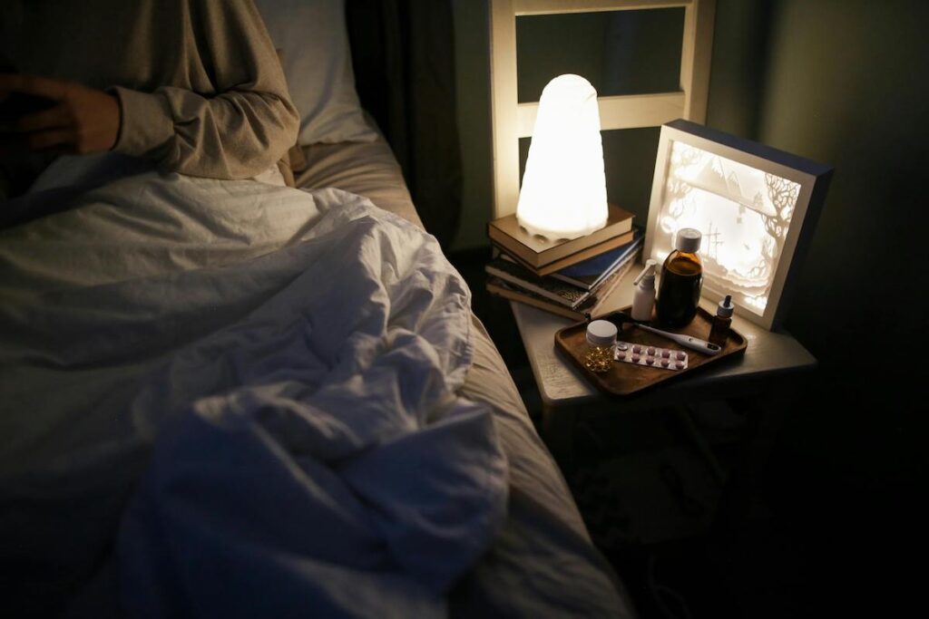 Night stand with a lamp and pills by the side of bed.