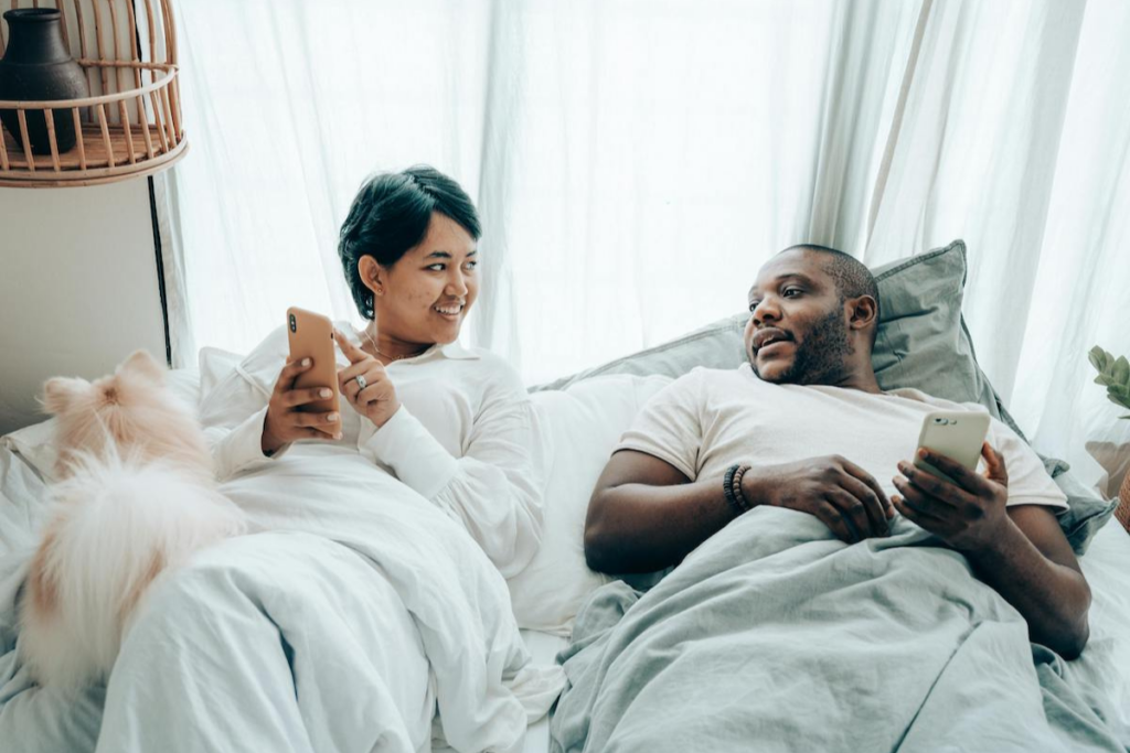 Two partners in bed looking at their phones and discussing.