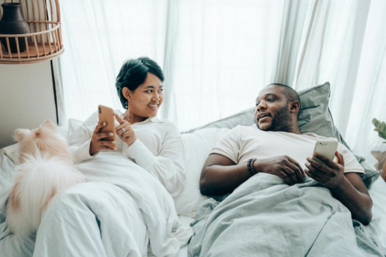 Two partners in bed looking at their phones and discussing.