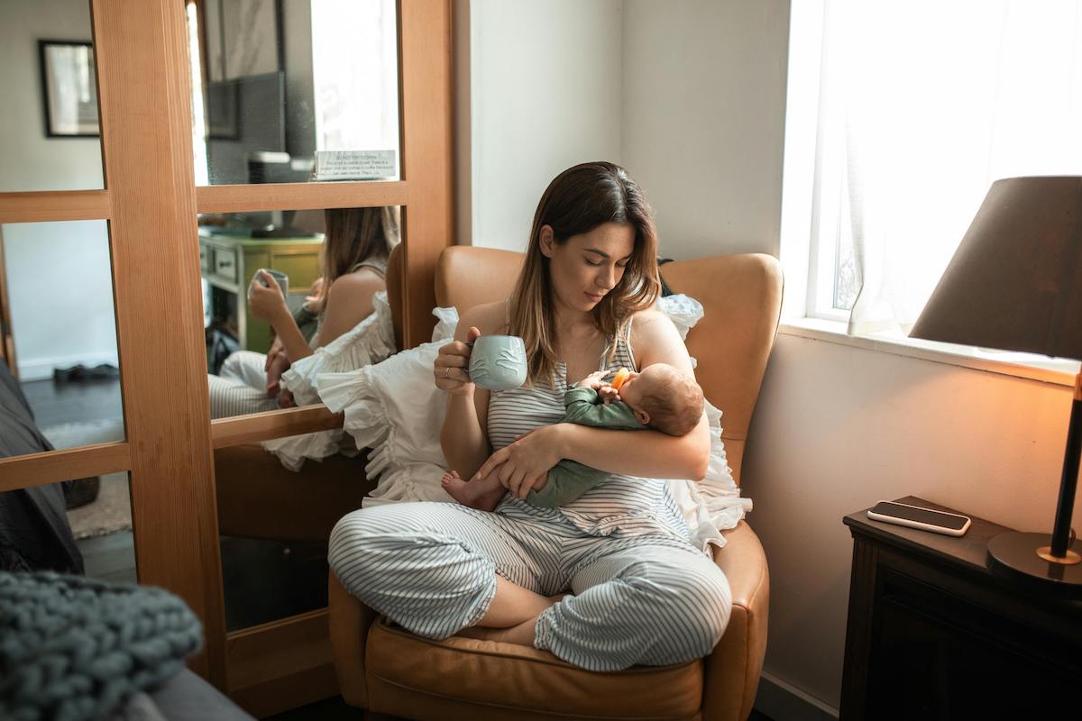 A mother sitting in a chair and drinking coffee while looking at the baby in hand.