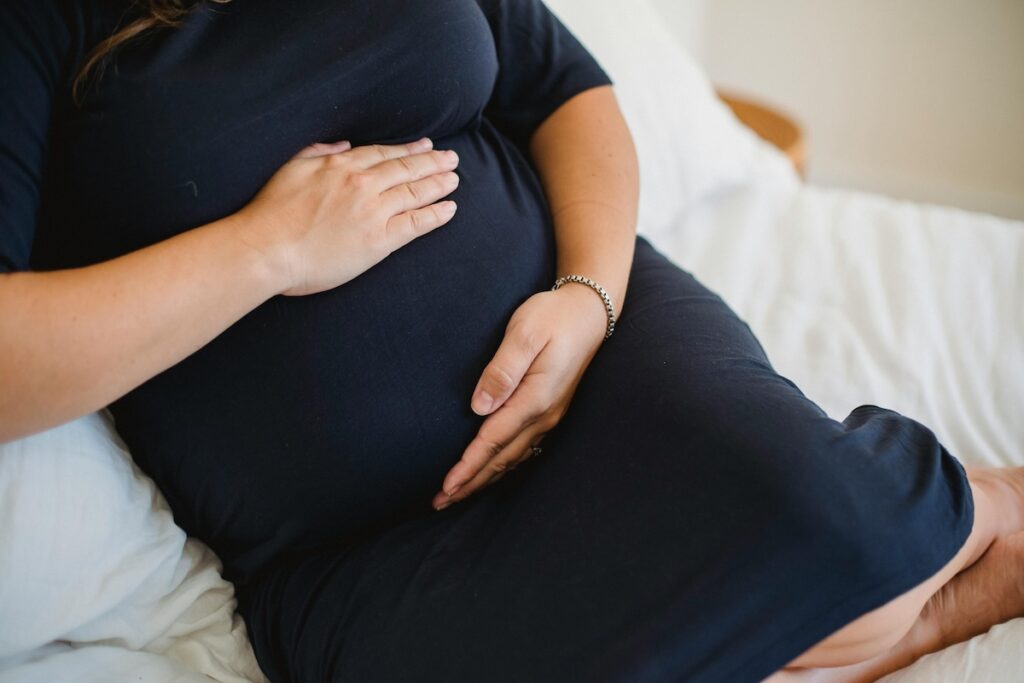 Pregnant person in a dark dress leaning against pillows in bed, with her hands on her stomach.